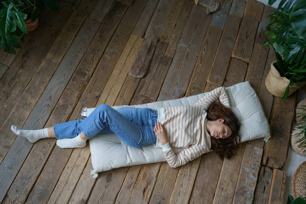 Donna italiana rilassata sdraiata sul pavimento in legno a casa, riposo dopo il lavoro. Vista dall'alto della calma giovane spagnola che dorme sul materasso circondata da piante tropicali in serra. Concetto benessere.  - Foto, immagini