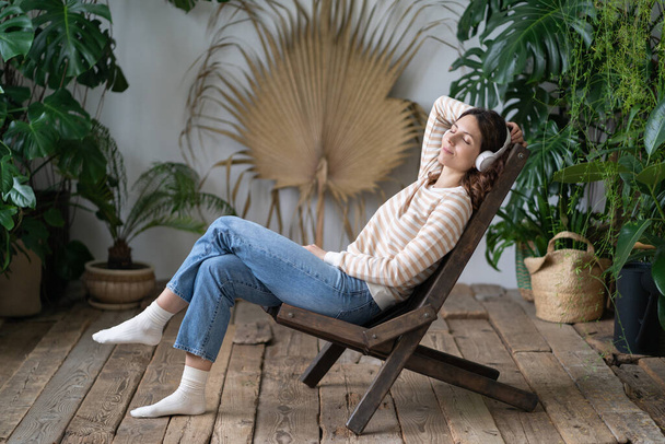 Спокойная женщина с закрытыми глазами в джинсах с беспроводными наушниками, отдыхающая в перерыве на работе, сидящая на уютном стуле в теплице с монстром, наслаждающаяся хорошей расслабляющей музыкой, дремлющая, мечтающая. Свободная от стресса концепция - Фото, изображение
