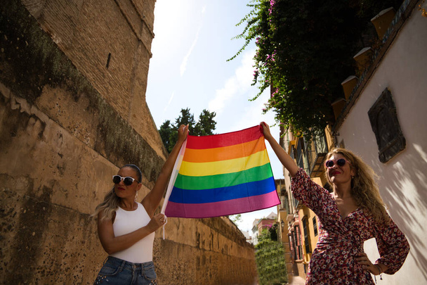duas mulheres bonitas usando óculos de sol seguram a bandeira do orgulho gay em sua mão. As mulheres estão felizes celebrando o dia do orgulho gay. Conceito de igualdade, homossexualidade, lgtb - Foto, Imagem