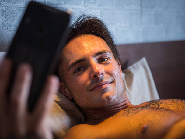 Attractive athletic young man using cell phone to take selfie photo or watching something on mobile, while laying shirtless on bed, shirtless - Фото, изображение