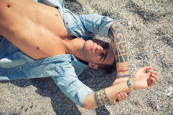 Stylish handsome young man laying on sand at the beach, shielding green eyes from the sunlight with his arm - Zdjęcie, obraz