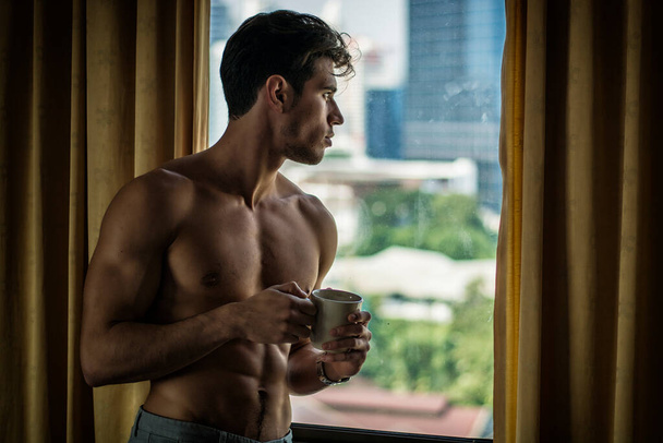 Sexy handsome young man standing shirtless in his bedroom drinking a cup of coffee or tea next to window curtains - Фото, изображение