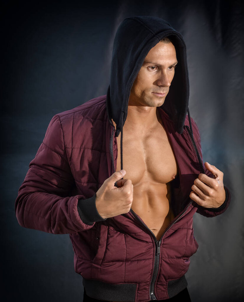 Muscleman opening his hoodie sweater revealing muscular torso, on dark background - Fotó, kép