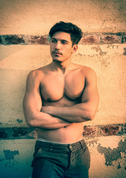 Atletische jongeman shirtloos in jeans voor betonnen muur buiten, met armen gekruist op zijn borst, tonen sexy lichaam, weg te kijken naar een kant - Foto, afbeelding