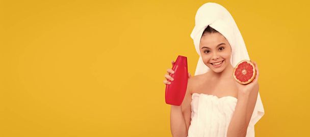 smiling kid in towel with grapefruit shampoo bottle on yellow background. Cosmetics and skin care for teenager child, poster design. Beauty kid girl banner with copy space - Foto, Bild
