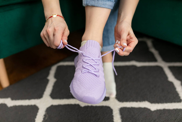 Cropped photo of woman wearing jeans, sitting on green sofa at home, lacing up violet sneakers, stretching laces, preparing for walking, running, sport, training workout. Sportswear, active lifestyle. - Zdjęcie, obraz