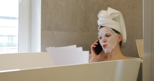 Worried annoyed young woman sitting in bath in bathroom with hair wrapped in towel, applying textile mask on face, talking arguing discussing on phone, holding sheets of paper. Relaxation, emotions. - Filmati, video