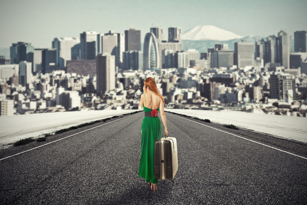 woman with suitcase walking on road towards city talking on phone - Valokuva, kuva