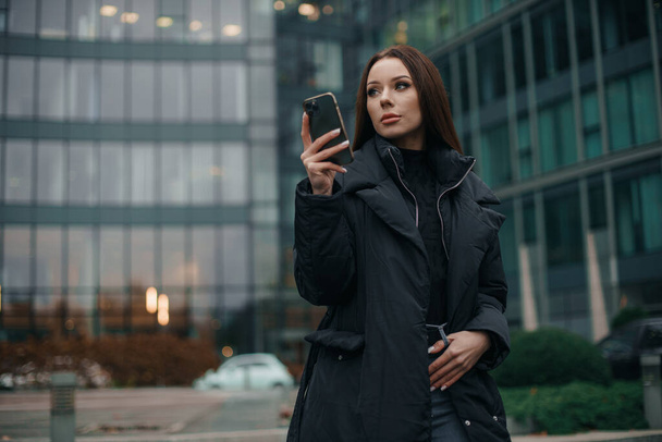Brunette girl with smartphone, standing on street of city centre, smiling at camera. Business woman holding cellphone using smartphone standing or walking on big city urban street outside. - Foto, afbeelding