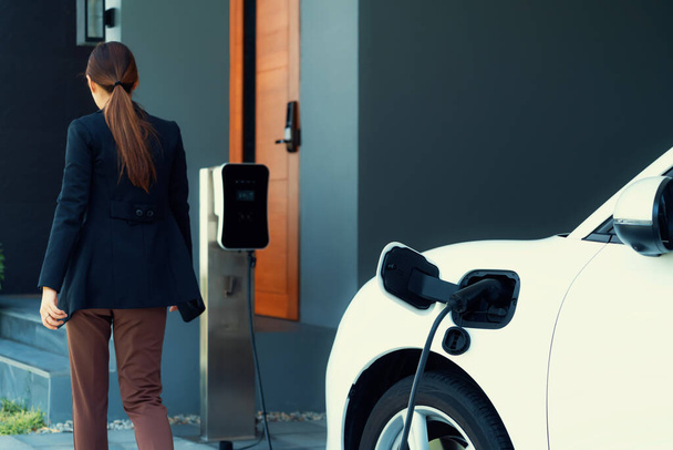 Progressive woman install cable plug to her electric car with home charging station. Concept of the use of electric vehicles in a progressive lifestyle contributes to clean environment. - 写真・画像