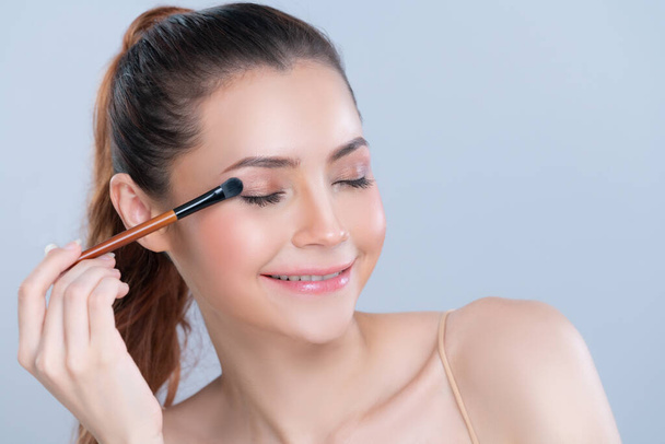 Closeup beautiful girl with flawless applying glamorous eye shadow makeup with eyeliner brush. Cosmetic facial painting process on lovely young woman with perfect clean skin in isolated background. - Foto, Imagen