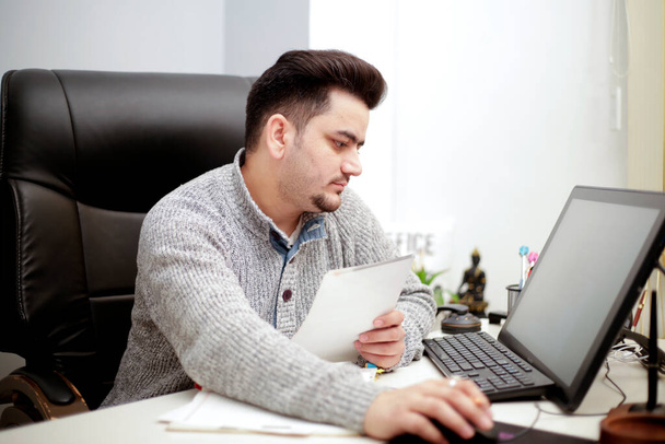Ein Geschäftsmann arbeitet am Computer, während er das Papier in der Hand hält. - Foto, Bild