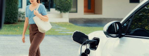 Progressive asian woman and electric car with home charging station. Concept of the use of electric vehicles in a progressive lifestyle contributes to a clean and healthy environment. - Zdjęcie, obraz
