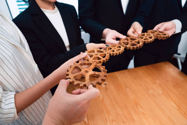 Closeup hand holding wooden gear by businesspeople wearing suit for harmony synergy in office workplace concept. Group of people hand making chain of gears into collective form for unity symbol. - Photo, Image