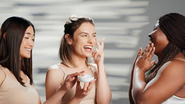 Multiethnic group of girls applying cream on face, using moisturizing cosmetics to prevent wrinkles. Positive happy women advertising bodycare campaign for skincare routine, different bodies. - Foto, imagen