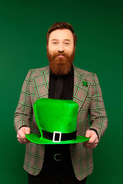 Smiling bearded man in jacket holding hat while celebrating saint patrick day isolated on green  - 写真・画像