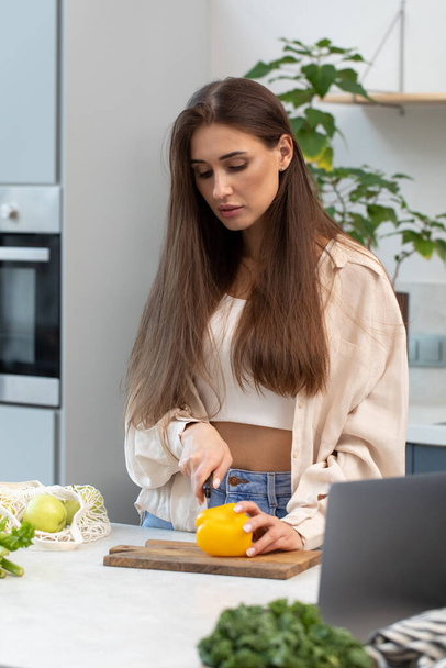 Attractive woman cuts vegetables for salad while looking at laptop screen. Video recipe. Healthy food. - Photo, image