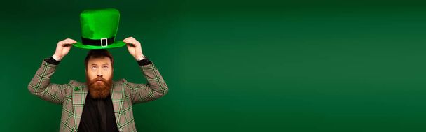 Bearded man in plaid jacket holding saint patrick day hat isolated on green, banner  - Fotoğraf, Görsel