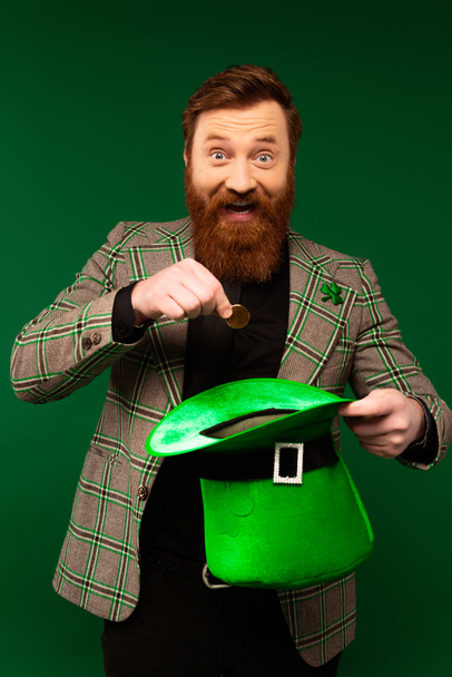 Homme barbu souriant regardant la caméra tout en tenant pièce de monnaie et chapeau isolé sur vert  - Photo, image