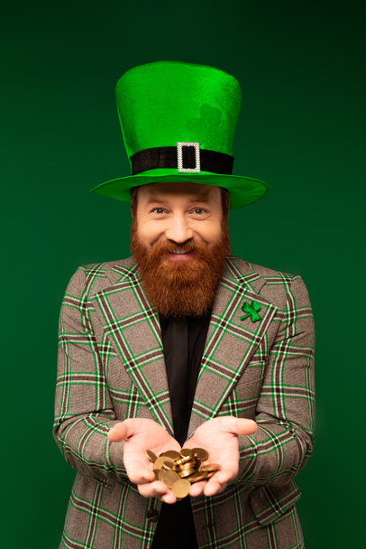 Smiling bearded man in hat holding coins while celebrating saint patrick day isolated on green  - Valokuva, kuva