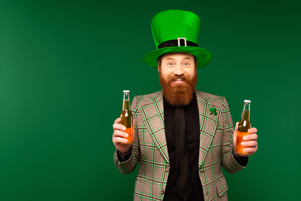 Smiling bearded man in hat holding bottles of beer isolated on green  - Foto, immagini