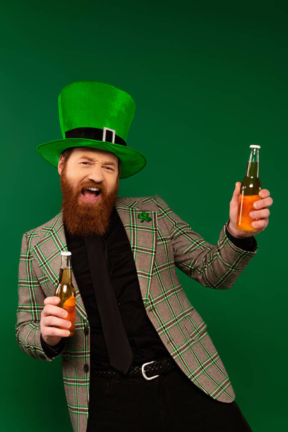 Excited man in jacket and hat holding bottles of beer isolated on green  - Photo, image