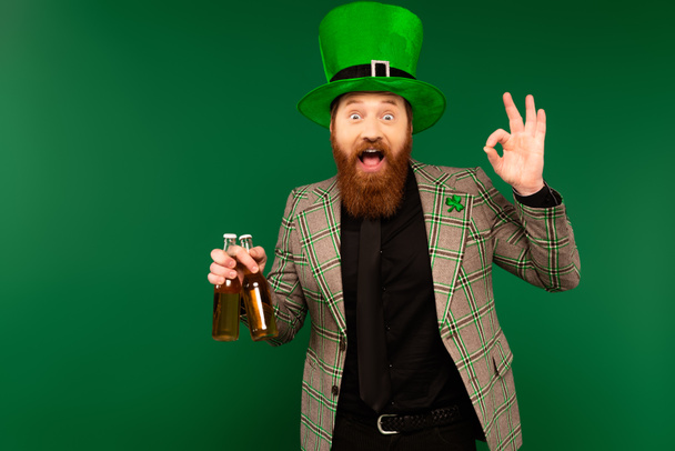 Excited man in hat holding bottles of beer and showing okay gesture isolated on green  - Photo, image
