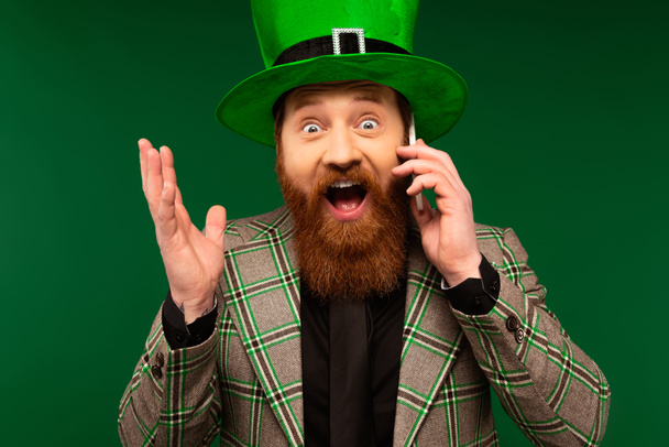 Astonished bearded man in hat talking on smartphone during saint patrick day isolated on green  - Photo, image