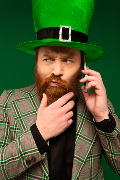 Thoughtful bearded man in hat with clover talking on smartphone isolated on green  - Fotografie, Obrázek