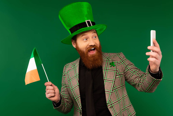 Smiling bearded man in hat holding Irish flag and taking selfie on smartphone isolated on green  - Fotó, kép