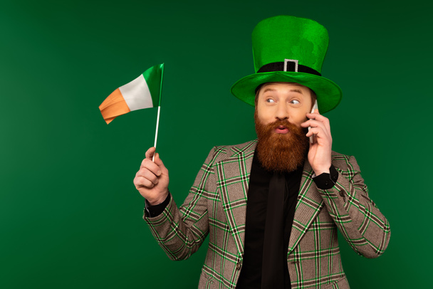 Bearded man in hat talking on smartphone and looking at Irish flag isolated on green  - 写真・画像