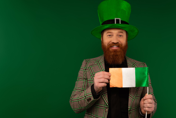 Un homme positif coiffé d'un chapeau tenant le drapeau irlandais pendant la fête de la Saint Patrick isolé sur vert  - Photo, image