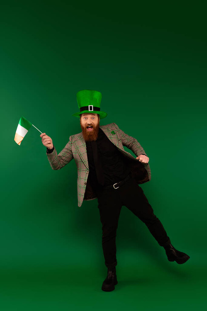 Πλήρες μήκος του χαρούμενου γενειοφόρος άνδρας στο καπέλο κρατώντας ιρλανδική σημαία σε πράσινο φόντο  - Φωτογραφία, εικόνα