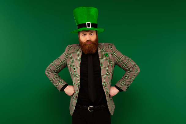 Злой бородатый мужчина в шляпе с клевером, держащимся за руки на бедрах, изолированных на зеленом  - Фото, изображение