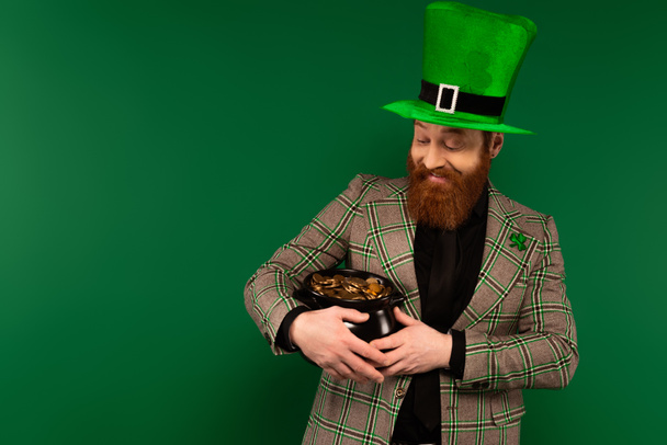 Smiling bearded man in hat holding pot with coins isolated on green  - Photo, Image