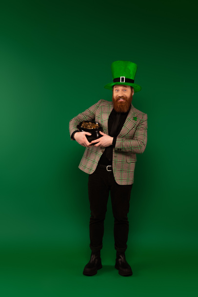 Cheerful bearded man in hat holding pot with golden coins on green background  - Photo, Image