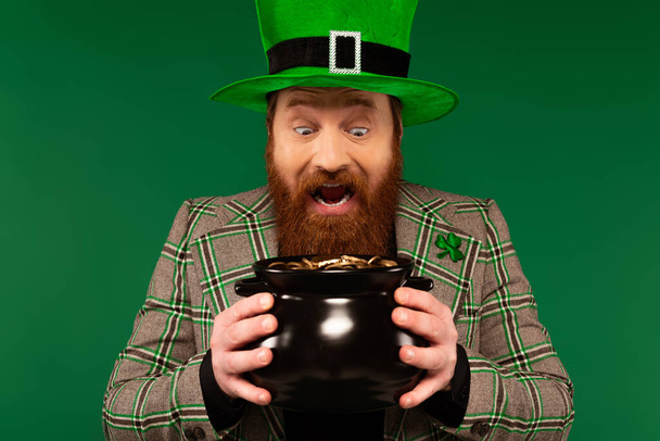 Hombre sorprendido en sombrero sosteniendo olla con monedas de oro aisladas en verde  - Foto, imagen