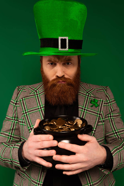 Θυμωμένος άνθρωπος στο καπέλο με τριφύλλι κρατώντας κατσαρόλα με χρυσά νομίσματα που απομονώνονται στο πράσινο  - Φωτογραφία, εικόνα