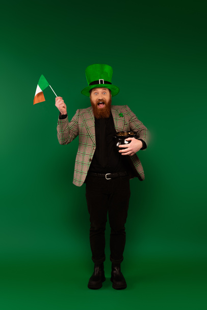 Full length of excited man in hat holding Irish flag and pot with coins on green background  - Photo, image
