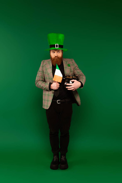 Pensive man in hat holding Irish flag and pot with coins on green background  - Foto, Imagen