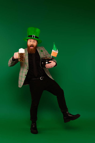 Excited man in hat holding beer and pot with coins and Irish flag on green background  - Foto, Imagen