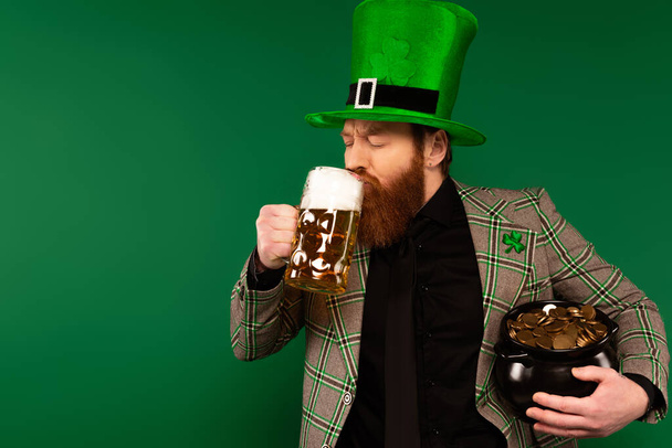 Bearded man in hat drinking beer and holding pot with coins isolated on green  - Foto, afbeelding