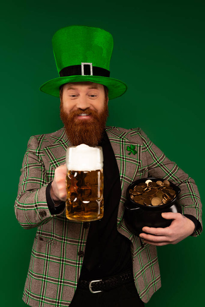 Веселый бородатый мужчина в шляпе держит стакан пива и горшок с монетами изолированы на зеленый  - Фото, изображение