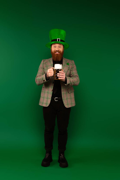 Πλήρες μήκος του χαμογελαστού γενειοφόρος άνδρας στο καπέλο κρατώντας ποτήρι μπύρα σε πράσινο φόντο  - Φωτογραφία, εικόνα