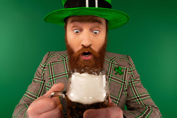 Hombre sorprendido en sombrero y trébol en chaqueta sosteniendo vidrio borroso de cerveza aislado en verde  - Foto, imagen