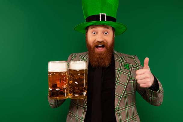 Ενθουσιασμένος άνδρας με καπέλο με τριφύλλι που δείχνει σαν και κρατώντας ποτήρια μπύρας που απομονώνονται σε πράσινο  - Φωτογραφία, εικόνα