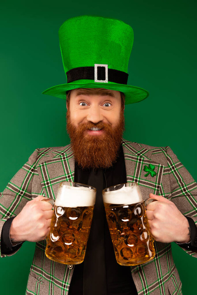 Uśmiechnięty mężczyzna w kapeluszu ze szklankami piwa odizolowanymi na zielono  - Zdjęcie, obraz