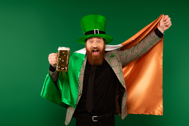 Возбужденный мужчина в шляпе, держащий стакан пива и ирландский флаг, изолированный на зеленом  - Фото, изображение