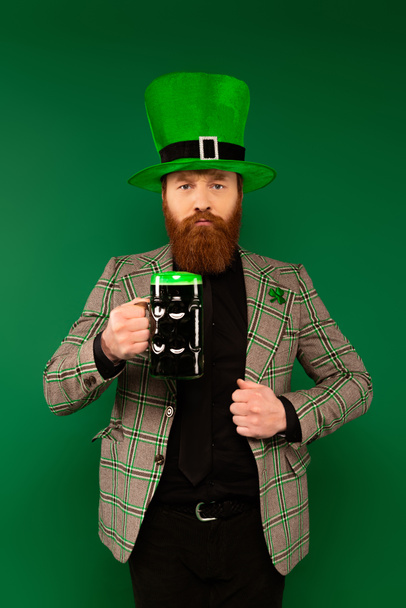 Hombre barbudo serio con sombrero sosteniendo un vaso de cerveza aislado en verde  - Foto, Imagen