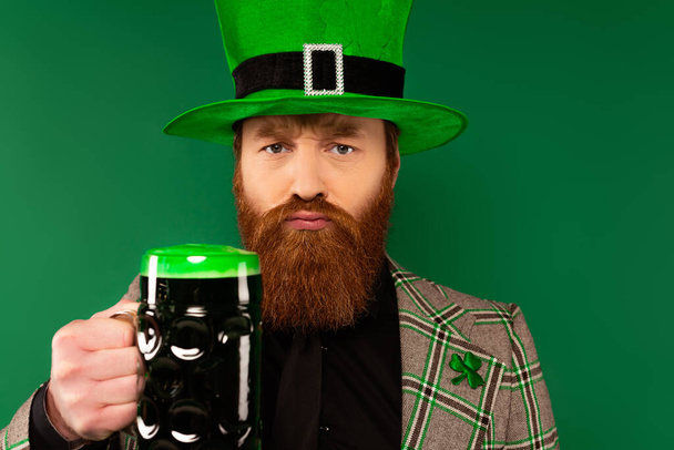 серьезный бородатый мужчина в шляпе держа стакан пива и глядя на камеру изолированы на зеленый  - Фото, изображение